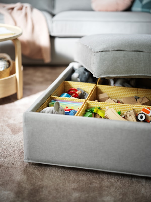 一个浅灰色KIVIK脚凳和存储盖子半价,揭示玩具分类在多个黄色UPPRYMD盒子。