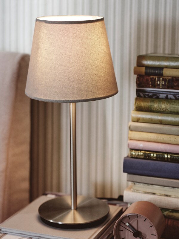 一个镀镍灯头SKAFTET表浅灰色SKOTTORP灯罩旁边小桌上一堆书。