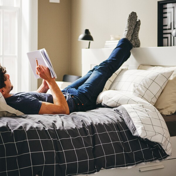 一个人躺在白色BRIMNES床上存储和床头板,用脚在床头板上,拿着书和铅笔。