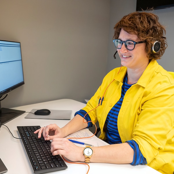 一名身穿蓝黄相间的宜家员工服、戴着眼镜和耳机的微笑女子坐在办公桌前看着亚博平台信誉怎么样屏幕。