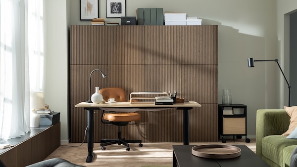 一个阳光充足的工作空间由BEKANT坐立两用桌和ALEFJÄLL办公椅组成，前面是一面墙的木质橱柜。