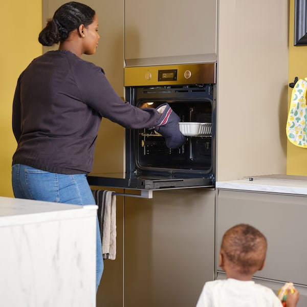 一个蹒跚学步的孩子看起来像一个女人需要烤箱菜的ANRATTA强迫通风烤箱在厨房马特黑色米色方面。