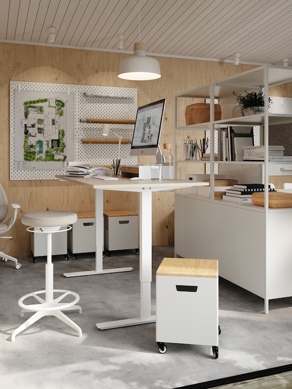 TROTTEN坐/立书桌，前面是LIDKULLEN主动坐/立支架，明亮的白色工作室带有开放式搁架。