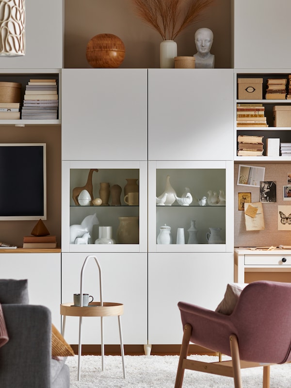 一个白色的BESTÅ存储系统展示装饰物品，墙上有一台电视，还有一把浅棕色的VEDBO扶手椅。