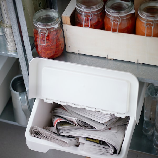 一个白色的SORTERA垃圾分类箱，打开盖子可以看到里面的报纸，下面是一架子的玻璃罐红色蜜饯。