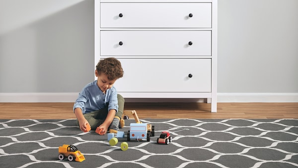 一个小孩玩大型玩具,有图案的地毯旁边白色HEMNES three-drawer胸部。