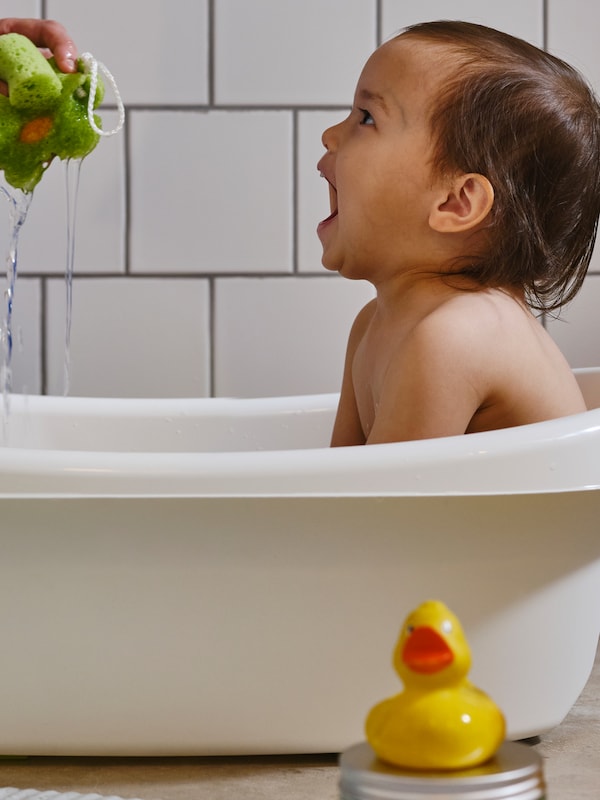 一个小孩坐在一个白色LATTSAM婴儿浴笑作为一个成年人的手挤压水从一层碧绿的海绵。