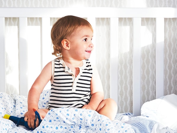 一个小孩坐在一个白色SUNDVIK床一边删除和GULSPARV床单与蓝莓的模式。