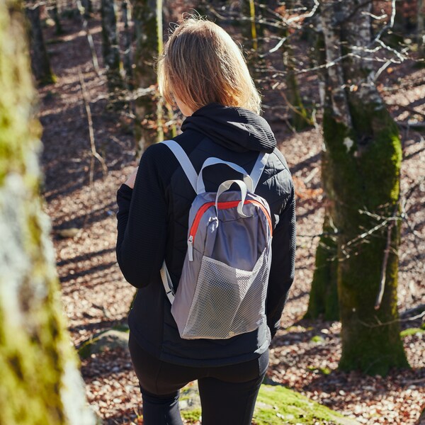 一个年轻人背着浅灰色的PIVRING背包，走在阳光明媚、森林稀疏的山坡下。