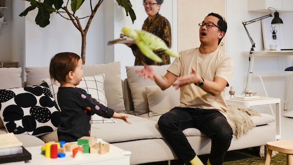 大人坐在浅肤色SODERHAMN沙发扔一个绿色软玩具一个挥舞着双臂站附近的孩子。