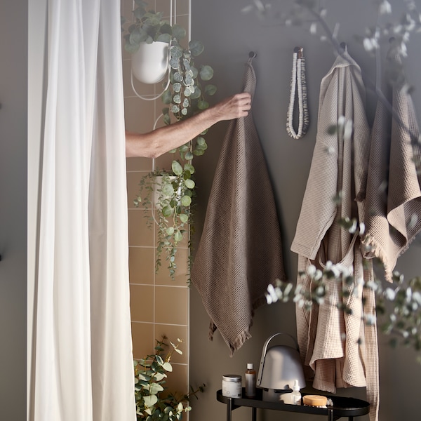 一只手臂从淋浴间伸出去拿挂在钩子上的浅灰色/棕色毛巾，外加浴帘和人造盆栽。