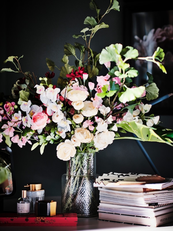 的安排SMYCKA人造花在透明玻璃/粉红色调图案KONSTFULL花瓶旁边一堆书。