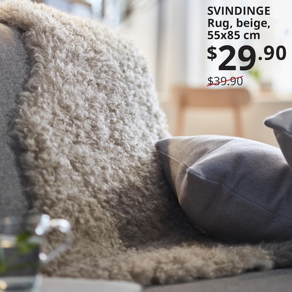 一个白色SVINDINGE地毯放在沙发上扔,和暗灰色坐垫放在地毯上。文字写着:SVINDINGE地毯,米黄色,x85 55厘米,29.90美元,之前的价格39.90美元。