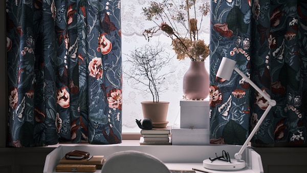 窗帘用深蓝色/花卉FILODENDRON织物在窗口上面白色与白色NYMANE HAUGA书桌灯工作。