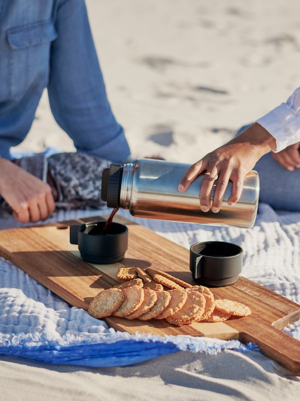 Dwie osoby siedząna kocu piknikowym, jedna z nich nalewa泽stalowego termosu UTRUSTNING kawę做czarnych kubkow wielokrotnego użytku。