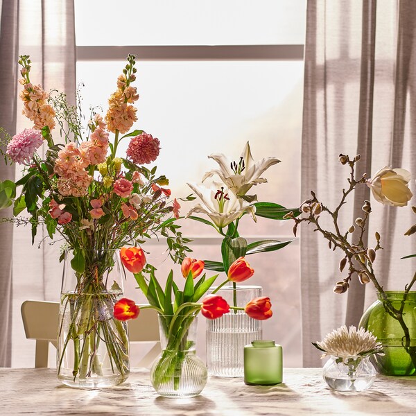 五个花瓶在不同大小和材料用鲜花,放在桌子上,旁边还有一个米色桌布一扇窗。