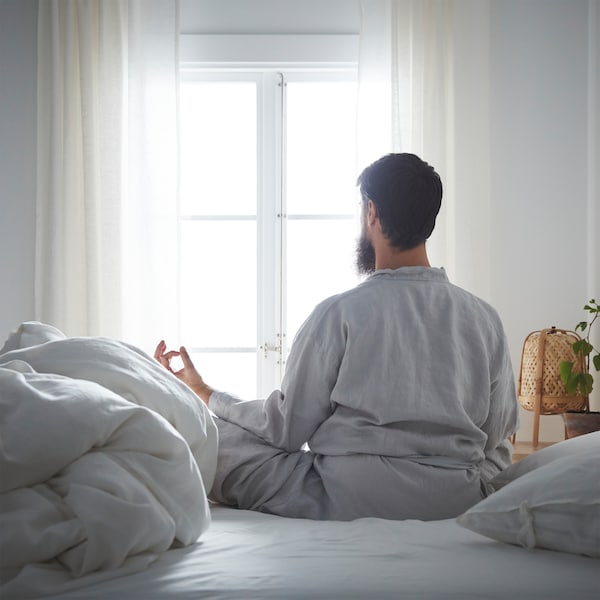 在calm-looking卧室,一个有胡子的人在冥想姿势盘腿坐在床上,看着窗外。