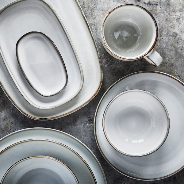 浅灰色的碗、盘子和餐具三堆，旁边的灰色表面上放着一个杯子。