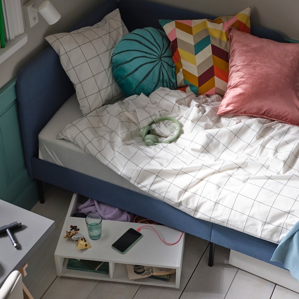 Modrá čalouněná postel s bílo-černým kostkovaným povlečením VITKLÖVER a různými polštáři, a s úložným prostorem pod postel FREDVANG。