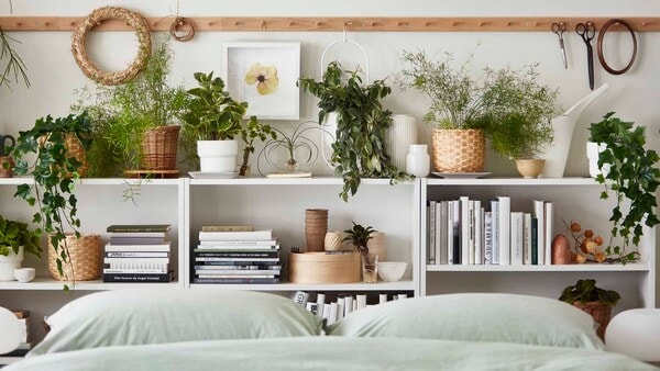 在一张铺有BERGPALM床上用品的床旁，书架上的书籍和装饰之间摆放着许多盆栽植物，它们都是真实的和人造的。