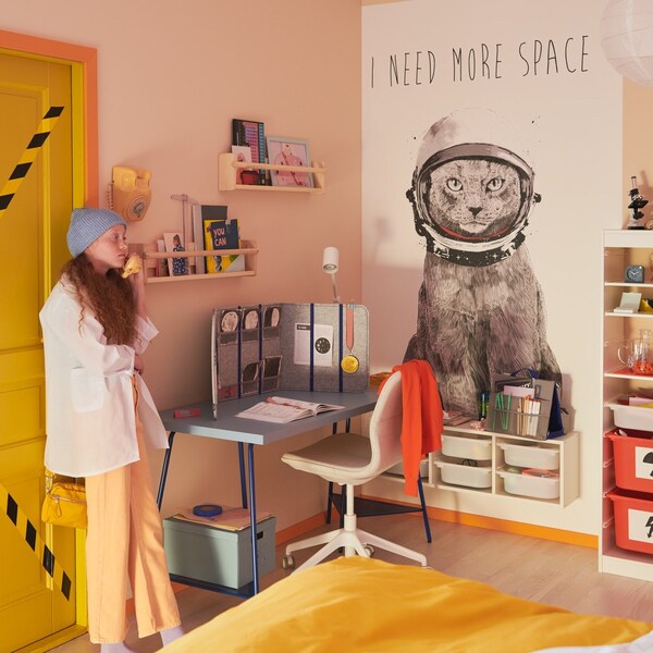Nastolatka stojąca wżołtych drzwiach swojego pokoju。Obok niej stoi biurko z fotelem biurowym一naścianie wisi duży plakat。