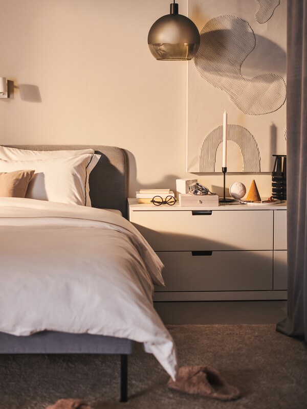 O lustrenko JAKOBSBYN montatska deasupra unei comode albe lângă un pat tapiat SLATTUM gri-deschis。