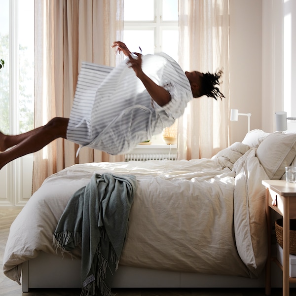 O persoanska care sare pe pe un pat MALM cu 2 cutii de depozitare într-un luminos cu draperii DYTÅG deschise la ferestre。