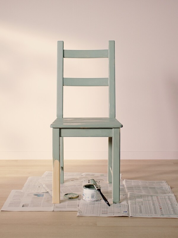 Pomalowane na jasnozielono krzesło IVAR ustawione na wyłożonej starymi gazetami jasnej, drewnianej podłodze w pomieszczeniu z jasnorożowymiścianami。