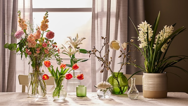 几个花瓶在不同大小和材料用鲜花,放在桌子上,旁边还有一个米色桌布一扇窗。