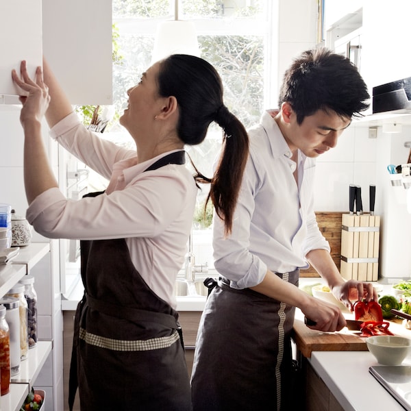 两个年轻人在一个小空间厨房围裙,一个是打开一个柜子,另一个是切菜。