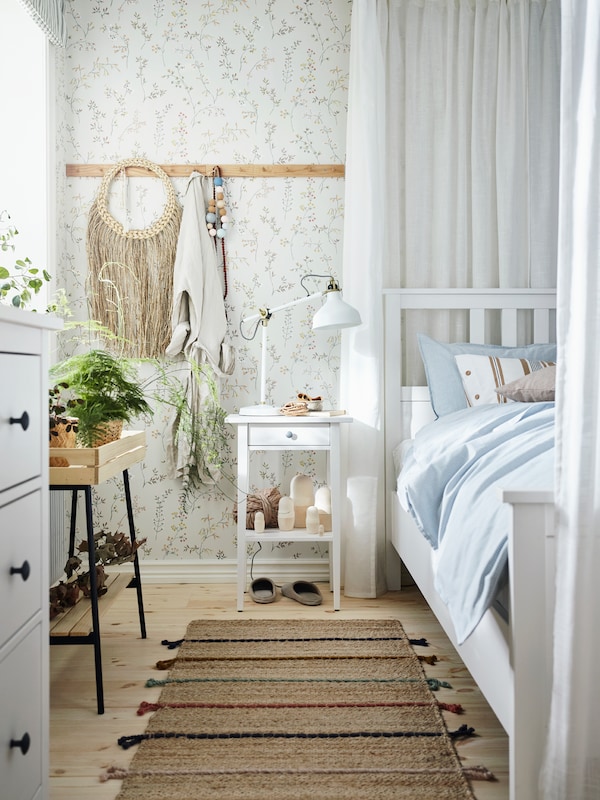 Un pat HEMNES alb lângă o noptierska pe care este azezatova o veiozova RANARP， într-un însorit, cu plante i decoraiuni naturale。