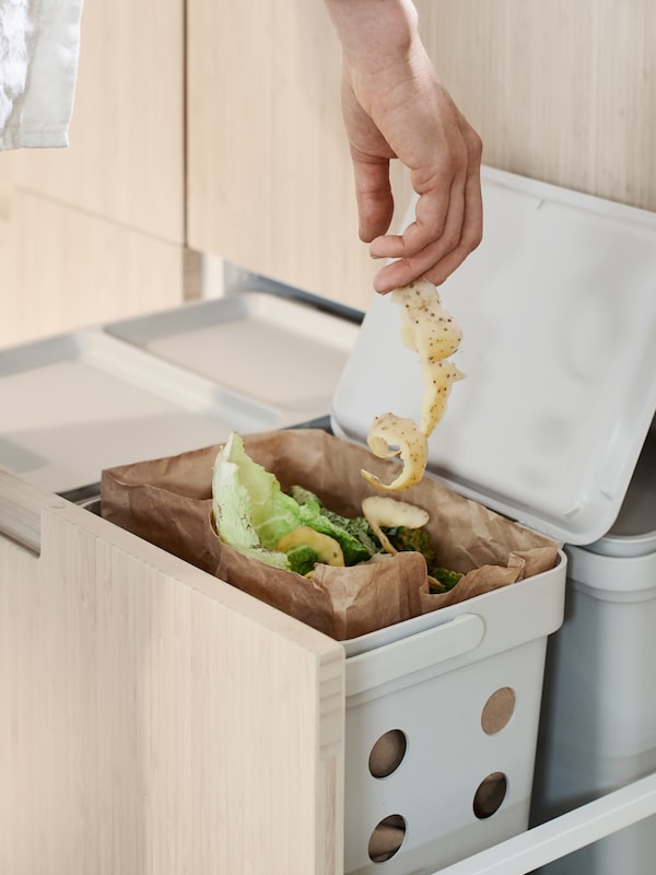 Une personne jetet Une épluchure de légume dans un sac à déchets alimentaires en paper brun。Cette dernière se trouve dan une solution de trii des déchets HÅLLBAR qui, elle, est installée dan un tiroir haut MAXIMERA。