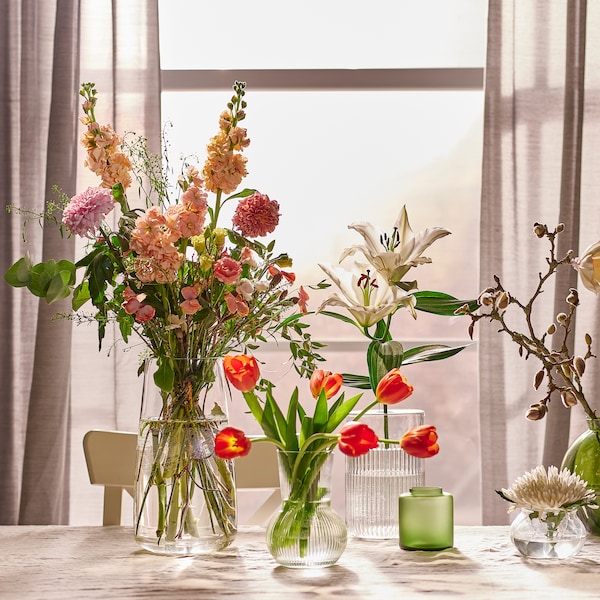 各种花卉安排绿色玻璃花瓶和透明玻璃花瓶在桌子上通过一个窗口灰色HILJA窗帘。