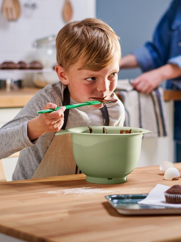 绿色/白色GUBBRORA抹刀,一个孩子在一个MARIATHERES围裙吃巧克力蛋糕面糊从绿色的碗里。